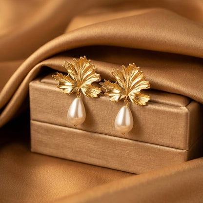 A pair of gold leaf-shaped drop earrings with teardrop pearl pendants, displayed on a gold jewelry box against a draped gold satin background.