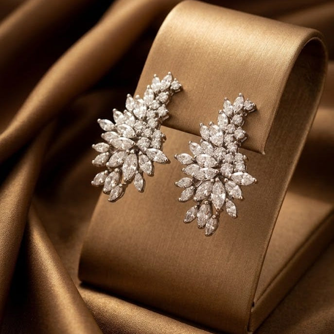 A pair of silver-toned leaf-shaped stud earrings set with clear stones, displayed on a curved champagne-colored jewelry stand against draped satin fabric.