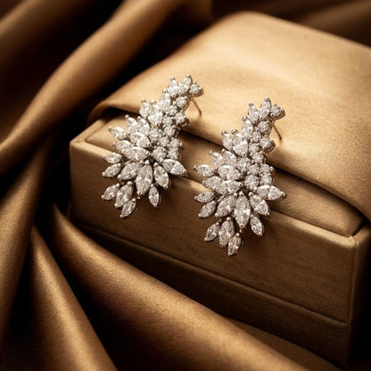 A pair of snowflake-shaped diamond earrings in a silver-tone setting, displayed on a gold velvet jewelry box against a draped brown satin background.