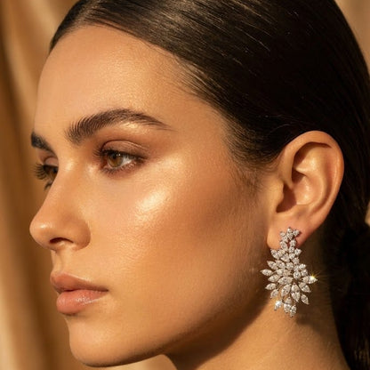 A close-up side profile of a woman with dark hair pulled back, wearing a sparkling silver-toned cluster earring with clear stones against a warm beige draped background.