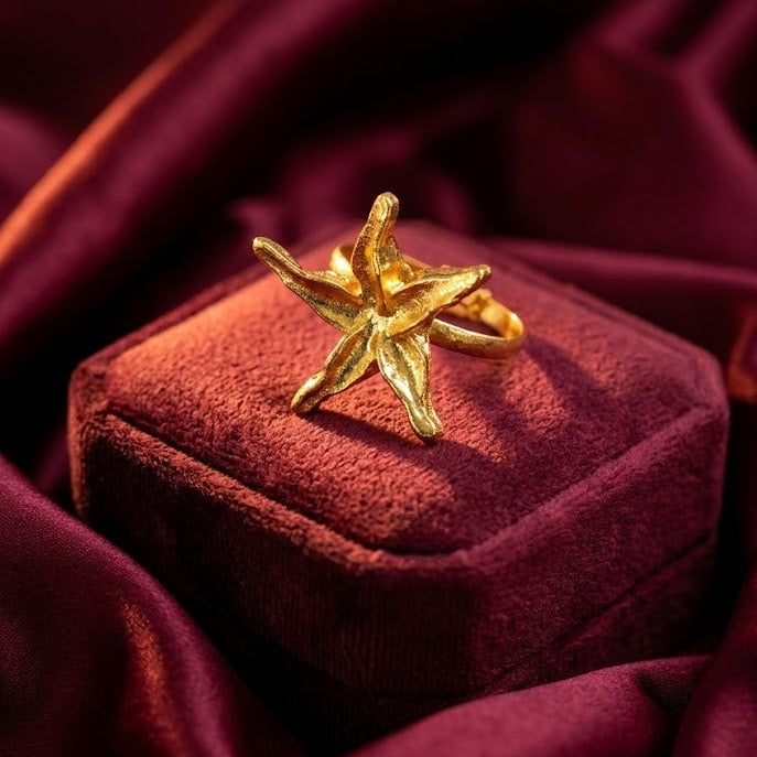 A gold starfish-shaped ring displayed on a textured burgundy velvet jewelry box, set against a backdrop of draped burgundy satin fabric.