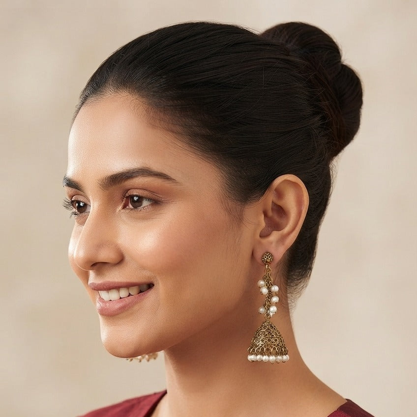 A woman with her hair in a bun is shown in profile wearing a deep maroon top and ornate gold-toned jhumka earrings with white pearl-like beads.