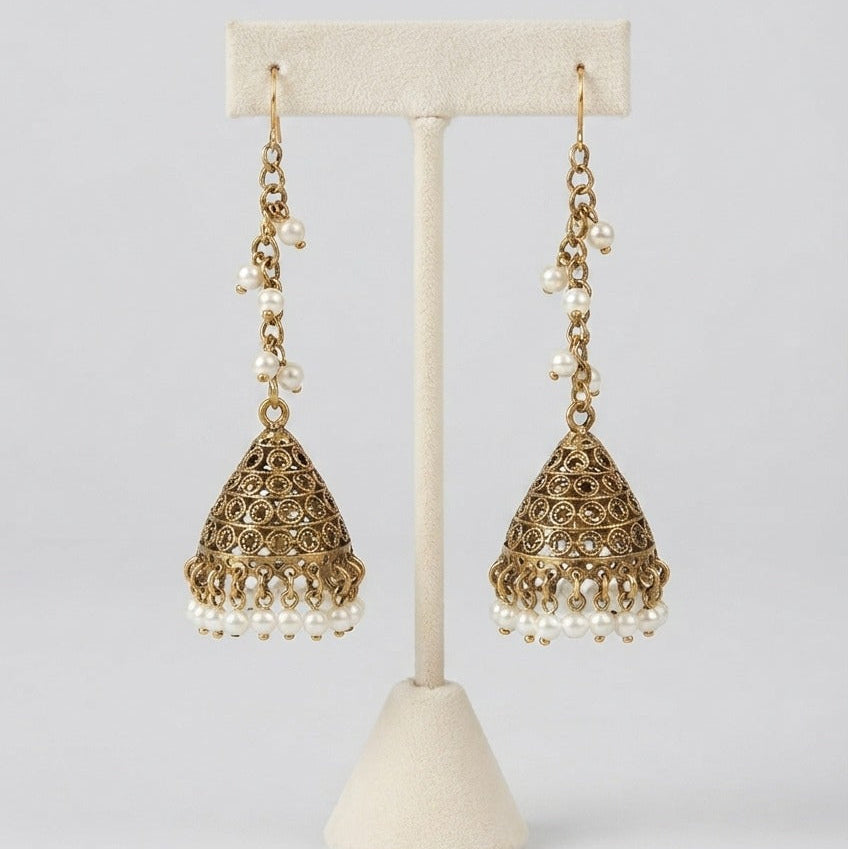 A pair of ornate gold-toned jhumka earrings with dangling pearl beads, displayed on a cream-colored jewelry stand against a light gray background.