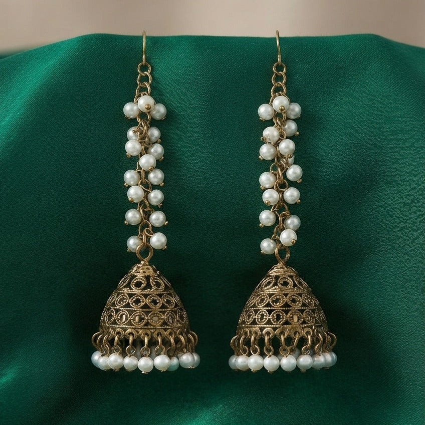 A pair of traditional Indian jhumka earrings with gold-toned filigree bells, white pearl bead strands, and small pearl drops, displayed on a draped green satin fabric.