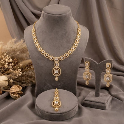 Gold jewelry set displayed on gray velvet mannequins, including a necklace, matching earrings, and a ring, arranged with dried floral decor against draped gray fabric.