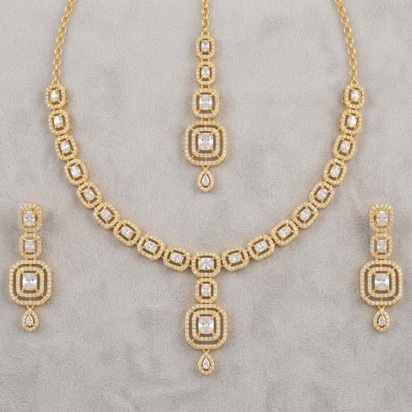 Gold jewelry set featuring a necklace, matching pendant, and two drop earrings, all adorned with clear stones in a geometric pattern, displayed on a textured gray background.