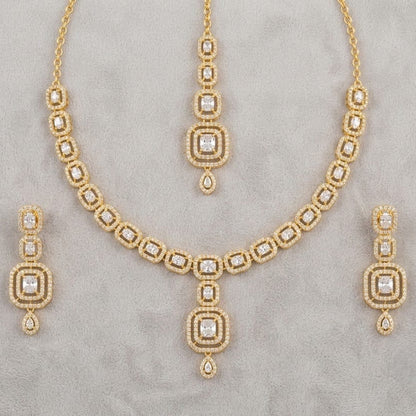 Gold jewelry set featuring a necklace, matching pendant, and two drop earrings, all adorned with clear stones in a geometric pattern, displayed on a textured gray background.