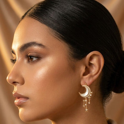A close-up side profile of a woman wearing a gold crescent moon dangle earring with small hanging charms, paired with a beige collared shirt against a warm beige background.