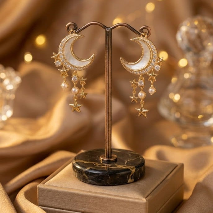 Gold crescent moon dangle earrings with pearl-like beads and star charms, displayed on a stand over a small jewelry box against a warm draped fabric background with soft bokeh lights.