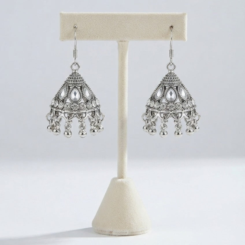 A pair of silver-toned traditional Indian jhumka earrings displayed on a cream-colored jewelry stand against a light gray background.