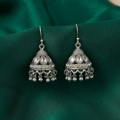 A pair of silver-toned Indian jhumka earrings with intricate filigree work and dangling bells, displayed on a draped emerald green satin fabric.