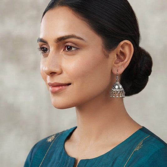 A woman in profile wearing a silver jhumka earring with a bell-shaped dangle and small hanging embellishments, paired with a teal embroidered top against a neutral textured background.