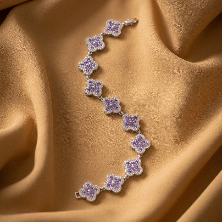 A delicate silver-toned bracelet with repeating lavender-purple flower motifs, laid out on draped champagne-gold fabric.