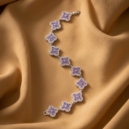 A delicate silver-toned bracelet with repeating lavender-purple flower motifs, laid out on draped champagne-gold fabric.