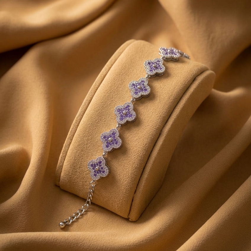 A delicate silver-toned bracelet with five small lavender-purple flower charms, displayed on a matching velvet jewelry stand against a draped tan fabric background.