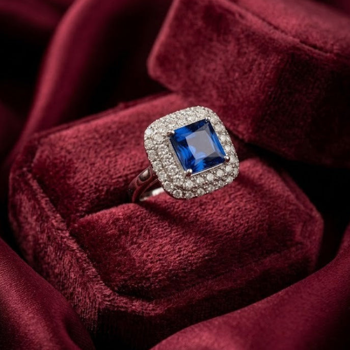 A square-cut blue gemstone ring with a diamond halo, displayed on a plush burgundy velvet ring box.
