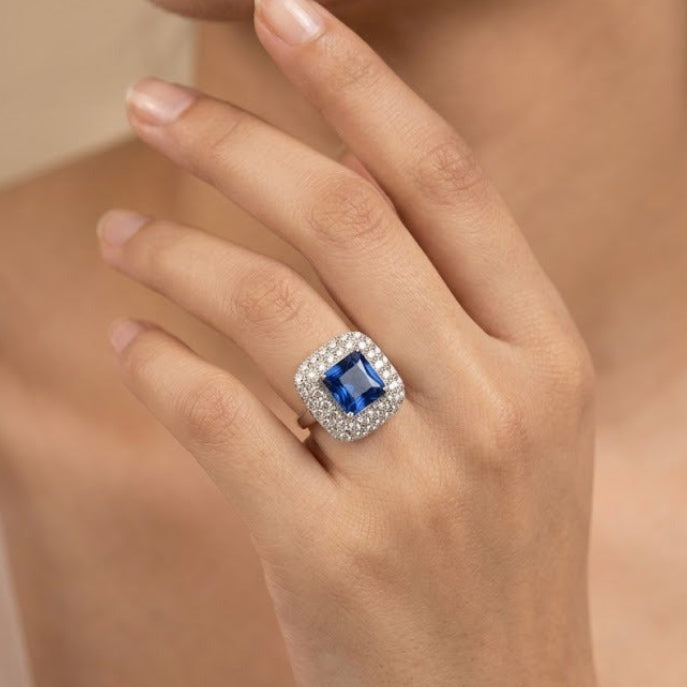 A close-up lifestyle shot of a model's hand wearing a ring with a square-cut blue gemstone surrounded by a halo of small clear stones in a silver-toned setting.