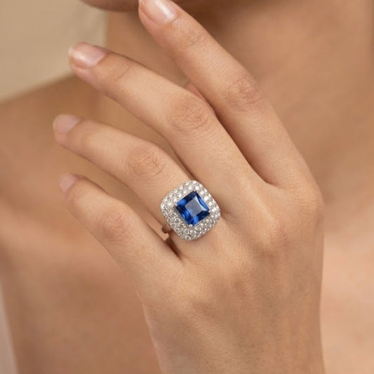 A close-up lifestyle shot of a model's hand wearing a ring with a square-cut blue gemstone surrounded by a halo of small clear stones in a silver-toned setting.