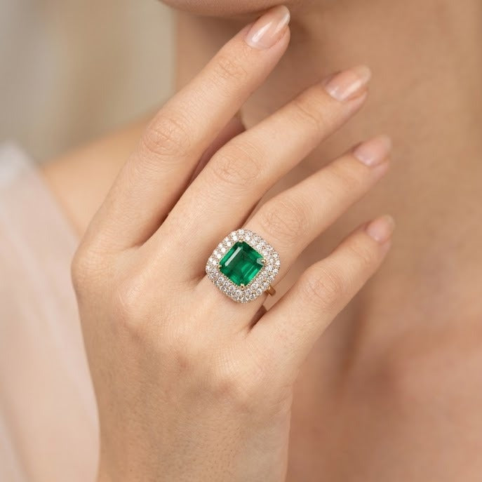 A close-up lifestyle shot of a model's hand wearing a luxury ring. The ring features a large cushion-cut emerald green center stone surrounded by a halo of small clear stones, set on a gold band.