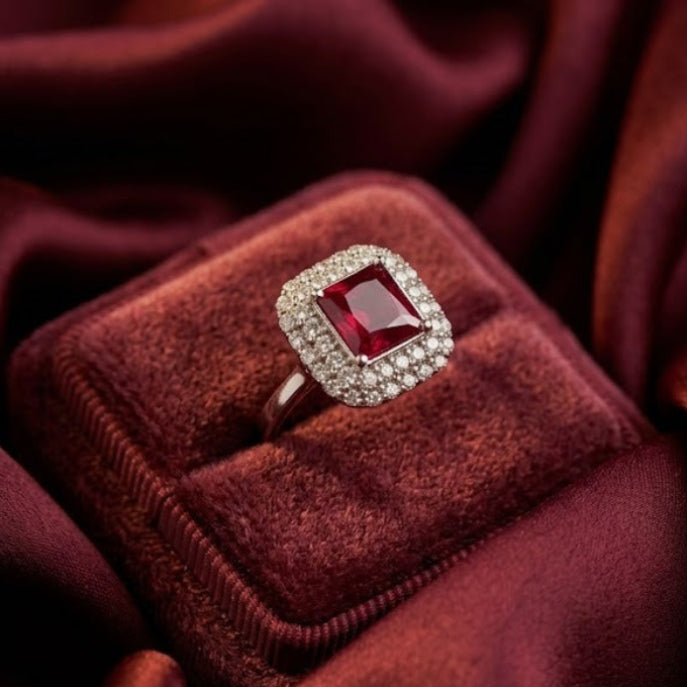 A square-cut red gemstone ring with a halo of small clear stones, displayed on a velvet ring box set against draped burgundy satin fabric.