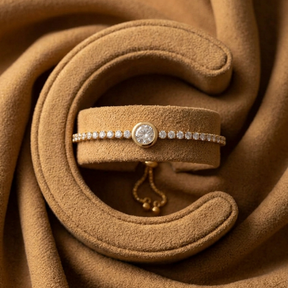 A delicate gold bracelet with a row of clear stones and a central round stone, displayed on a matching tan velvet jewelry stand against a draped tan fabric background.