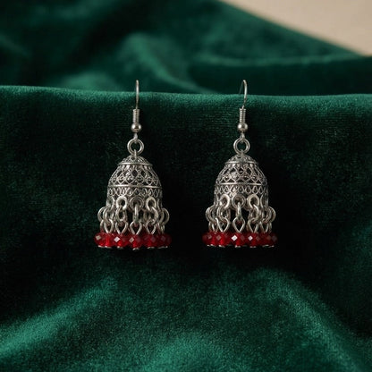 A pair of silver-toned filigree jhumka earrings with small red bead drops, displayed on a dark green velvet fabric background.