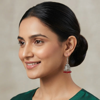 A woman with her hair in a bun wearing a dark green embroidered kurta and a silver-toned jhumka earring with red beads, shown against a neutral beige background.