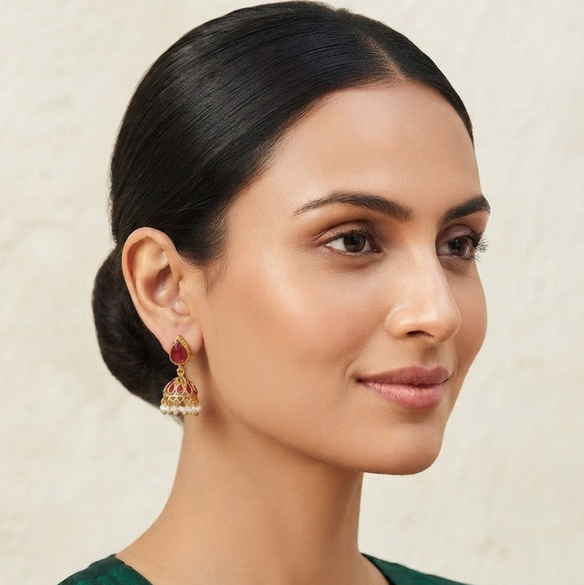A woman with her hair neatly tied back in a bun, wearing a dark green kurta with gold embroidery and a red jhumka earring, set against a textured beige wall.