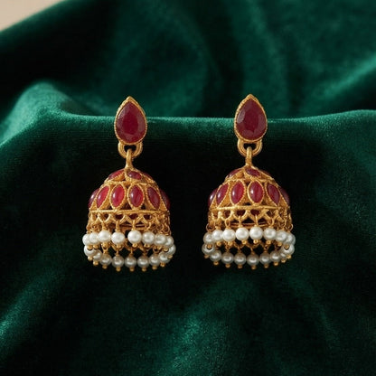 A pair of traditional Indian gold jhumka earrings with red stones, white pearls, and gold filigree, displayed on a dark green velvet fabric background.