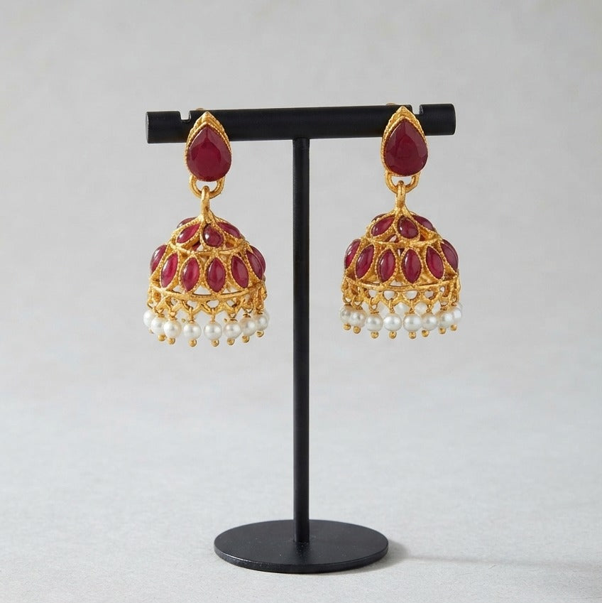 A pair of gold-plated Indian jhumka earrings with deep red enamel and white pearl bead drops, displayed on a black stand against a light gray background.