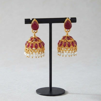 A pair of gold-plated Indian jhumka earrings with deep red enamel and white pearl bead drops, displayed on a black stand against a light gray background.