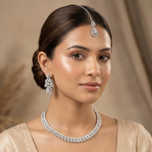 A woman wearing a silver-toned traditional jewelry set including a maang tikka, earrings, and necklace, paired with a beige traditional outfit against a soft beige draped background.