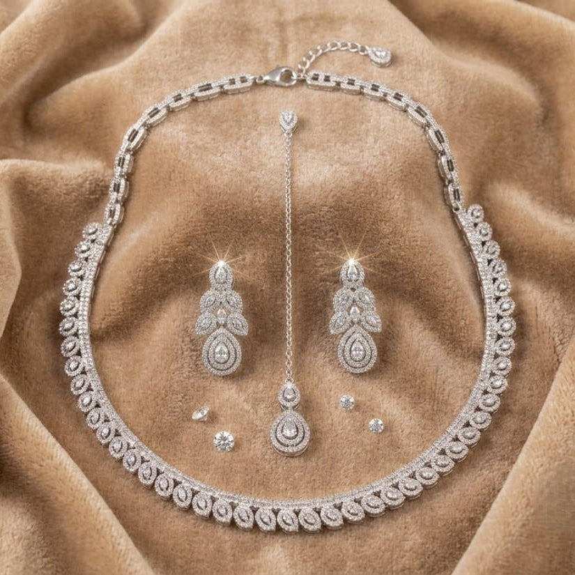 A silver-toned jewelry set featuring a necklace, matching earrings, and a long pendant, all adorned with sparkling clear stones, displayed on a draped tan velvet fabric background.