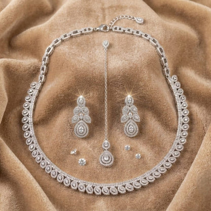 A silver-toned jewelry set featuring a necklace, matching earrings, and a long pendant, all adorned with sparkling clear stones, displayed on a draped tan velvet fabric background.