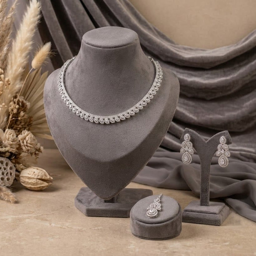Silver-toned jewelry set displayed on gray velvet stands, including a necklace, earrings, and ring, arranged with dried botanical decor and draped fabric in a styled studio setting.