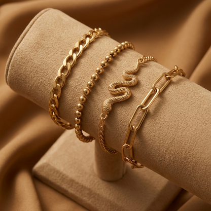 Four gold-toned bracelets displayed on a beige velvet jewelry stand, set against a draped tan fabric background. The bracelets include a curb chain, a beaded chain, a rope chain with a snake motif, and a paperclip chain.