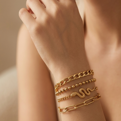A close-up of a person's wrist wearing a stack of gold bracelets, including a chunky chain link bracelet, a beaded bracelet, a snake-shaped bracelet, and a paperclip-style bracelet.