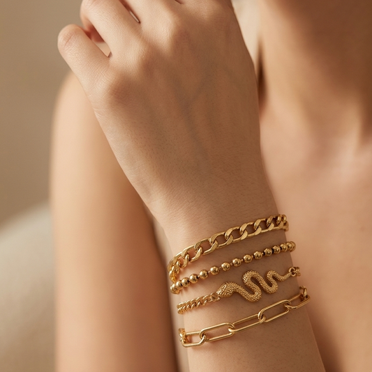 A close-up of a person's wrist wearing a stack of gold bracelets, including a chunky chain link bracelet, a beaded bracelet, a snake-shaped bracelet, and a paperclip-style bracelet.