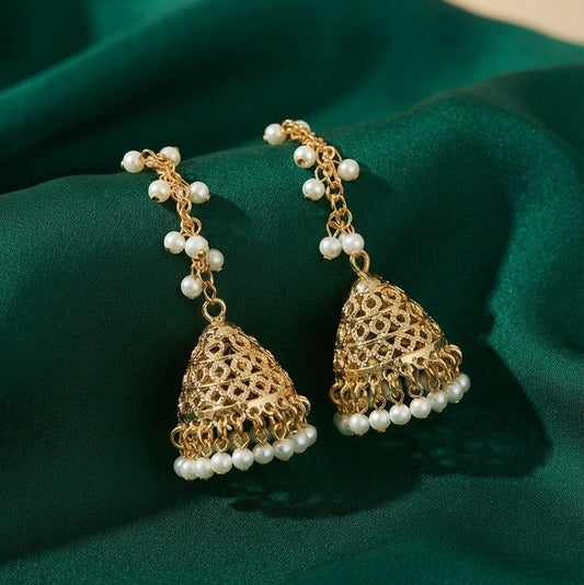 A pair of gold-toned jhumka earrings with white pearl bead accents, displayed on a draped dark green satin fabric.
