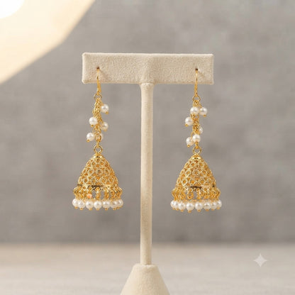 A pair of gold-toned jhumka earrings with white pearl bead accents, displayed on a cream-colored jewelry stand against a soft gray background.