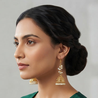 A woman in profile wearing a dark green embroidered kurta and gold-toned jhumka earrings with white pearl-like beads, set against a soft light gray background.