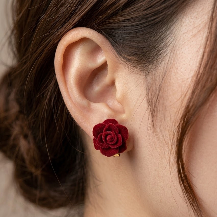 A close-up of a woman's ear wearing a small, deep red rose-shaped stud earring with a tiny gold accent at the base.