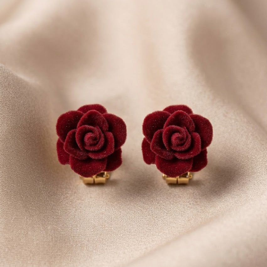 A pair of deep red velvet rose stud earrings with gold-tone posts, displayed on a soft beige satin fabric background.