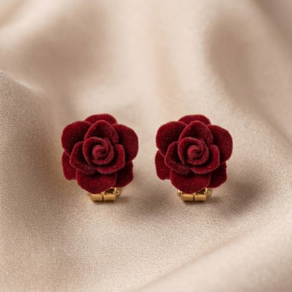 A pair of deep red velvet rose stud earrings with gold-tone posts, displayed on a soft beige satin fabric background.