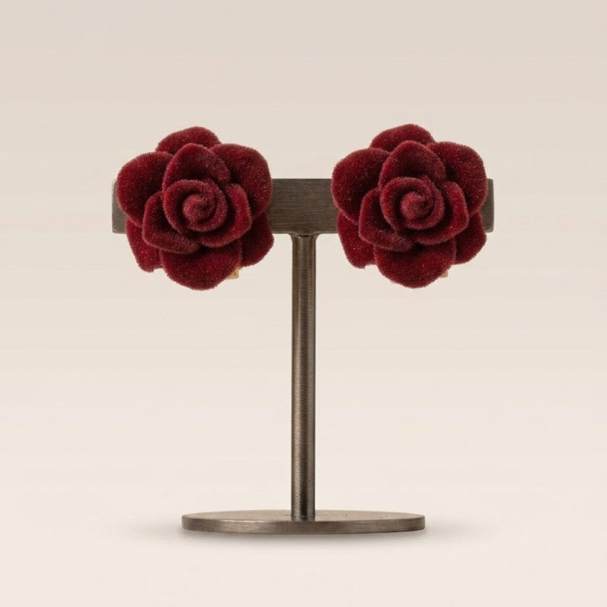 A pair of deep burgundy velvet rose stud earrings displayed on a dark metal stand against a warm beige background.