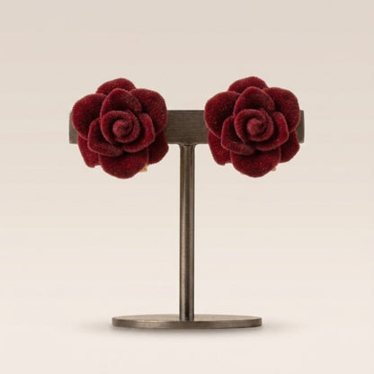 A pair of deep burgundy velvet rose stud earrings displayed on a dark metal stand against a warm beige background.