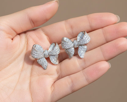 Three silver bow-shaped earrings held in a hand against a neutral background