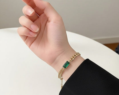 Hand wearing a gold bracelet with a green gemstone on a neutral background