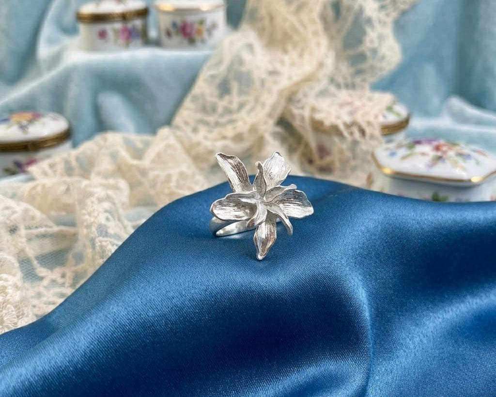Silver floral ring on blue satin fabric with lace and decorative boxes in the background