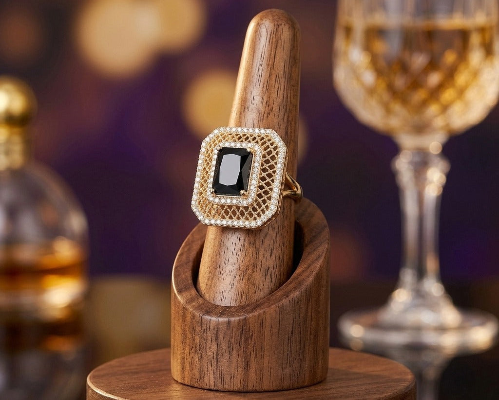Gold ring with a black gemstone on a wooden stand, with a blurred background of a glass and bottle.